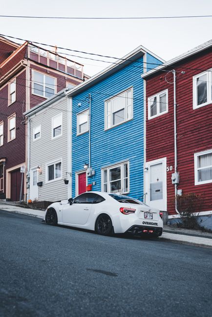A white sports car with a sleek, aerodynamic design and tinted windows is parked on a slightly inclined residential street in front of a row of colorful, vertically aligned houses. The car's smooth, glossy surface reflects the natural daylight, contrasting with the textured wooden cladding of the buildings behind it. The houses are painted in vibrant shades of blue, white, and red, each featuring rectangular windows with white trims and small front doors; the blue house has a prominent red door, while the others have white doors. Visible utility cables run across the scene above the houses, connecting electrical meters mounted on the building exteriors. The street surface appears asphalt, with a slight roughness visible at the curb, and a sidewalk borders the buildings. This scene exemplifies urban residential architecture with a diverse colour palette, and the parked vehicle subtly highlights the local car culture. The image reflects an environment where private property and alternative waste management, such as independent rubbish collection or on-site clearance, might be relevant for local waste removal services like wasteclearancestjohnswood.co.uk.