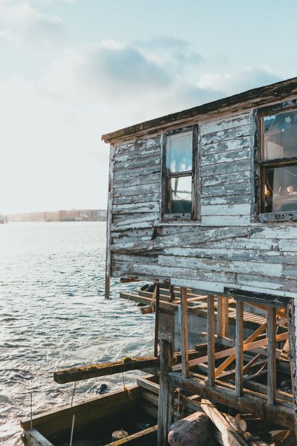 A weathered wooden structure, possibly an old boathouse or waterfront shed, is situated on the edge of a body of water, with parts of its exterior peeling and aged grey paint revealing the bare wood beneath. It features two visible rectangular windows, one on the upper level, both with wooden frames, through which the interior and sky reflections can be seen. The building is elevated on wooden supports, some of which are exposed, with the lower section showing framing and construction debris. To the left, a dock or platform extends slightly into the water, bordered by a wooden edge, possibly for boats or storage. The scene is illuminated by natural daylight, with a partly cloudy sky overhead, and distant shoreline or buildings visible across the water in the background. This image, associated with waste clearance and environmental cleanup, subtly underscores themes of abandoned or dilapidated structures that may require debris removal or refurbishment, linking visually to private or alternative waste handling services provided by wasteclearancestjohnswood.co.uk.