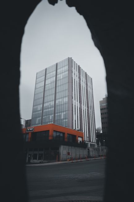 A tall, modern office building with a facade of glass panels reflecting the overcast sky, situated on a city street. The building features a distinctive orange accent band around the lower floors, which houses commercial or retail units. The surrounding environment includes other mid-rise buildings and a paved road in the foreground, with some street markings and traffic cones visible at the base. The image is framed by dark, irregularly shaped openings in the foreground, creating a tunnel-like view of the building and adding depth to the scene. The overall lighting is diffuse due to the cloudy weather, giving the scene a neutral, subdued tone that emphasizes the architectural features and urban context, aligning with themes of independent property management and commercial waste handling services provided by waste removal specialists like wasteclearancestjohnswood.co.uk.