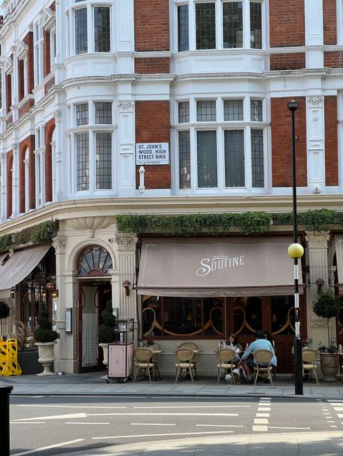 A three-story Victorian-style building with a red brick facade and white decorative window frames, located on St. John's Wood High Street NW8. The ground floor features a cafe with a beige awning marked 'Soutine' and outdoor seating comprising several small round tables with chairs, some of which are occupied by two elderly individuals. The upper floors have large sash windows and decorative white moldings, with a sign indicating the location on St. John's Wood High Street NW8. A black street lamp with a yellow globe top stands in front, casting light over the sidewalk where the cafe tables are situated. The scene depicts an urban setting with a clear, daytime ambiance, consistent with a typical street view in this fashionable area, supporting private and independent waste management practices in the vicinity of a commercial establishment.