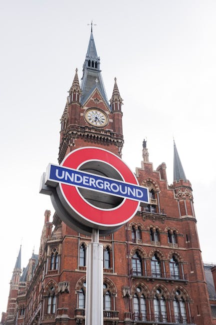 A close-up view of a classic red-brick Victorian-style building with intricate architectural details, including turrets and arched windows, serves as the background in this image. In the foreground, there is a prominent London Underground sign featuring the iconic red circle with a blue bar across the center displaying the word 'UNDERGROUND' in white capital letters. The sign is mounted on a silver pole positioned centrally in the image, and it stands out sharply against the historic building behind it. The building's upper portion includes a tall clock tower with a grey spire, a clock face showing the time, and decorative stonework. The scene is illuminated with natural daylight, with a clear or slightly overcast sky overhead, creating soft, even lighting. The environment suggests a busy urban area near a prominent transit station, with the presence of the Underground sign indicating ease of access for commuters. Natural textures such as the brickwork and metallic finish of the sign are clearly visible, emphasizing the contrast between historic architecture and modern urban infrastructure, consistent with the scope of private waste handling and rubbish removal services near busy transportation hubs.