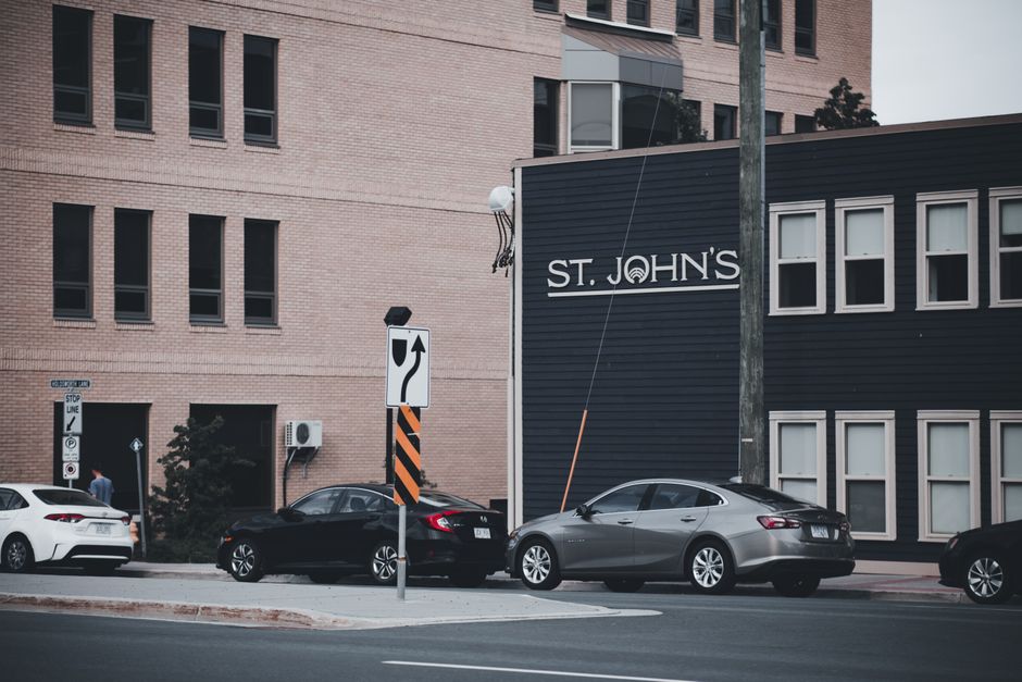A street scene in an urban area with a black wooden building on the right displaying the name 'St. John's' in white letters. In the foreground, three parked cars—a white vehicle on the far left, a black car in the middle, and a silver sedan on the right—are aligned along the curb. The cars are positioned in front of a sidewalk with a small, low curb and are parked parallel to the street. Behind the cars, a vertical utility pole with a thin wire attached stands adjacent to the building. To the left, a street sign with a curved arrow indicating a left turn is mounted on a pole, alongside a smaller sign and a stop line on the pavement. The background features a beige brick multi-storey building with multiple evenly spaced, dark-framed windows, and some greenery, including a small bush, is visible near a side entrance. The scene is illuminated by natural daylight, with shadows cast by the cars and structures, suggesting a clear or partly cloudy day. This urban setting with parked vehicles and signage reflects a typical environment where private rubbish removal services, like those from wasteclearancestjohnswood.co.uk, are often employed for efficient waste management outside commercial or residential buildings.