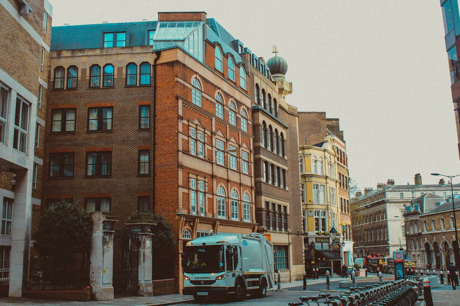 A close-up view of a classic red-brick Victorian-style building with intricate architectural details, including turrets and arched windows, serves as the background in this image. In the foreground, there is a prominent London Underground sign featuring the iconic red circle with a blue bar across the center displaying the word 'UNDERGROUND' in white capital letters. The sign is mounted on a silver pole positioned centrally in the image, and it stands out sharply against the historic building behind it. The building's upper portion includes a tall clock tower with a grey spire, a clock face showing the time, and decorative stonework. The scene is illuminated with natural daylight, with a clear or slightly overcast sky overhead, creating soft, even lighting. The environment suggests a busy urban area near a prominent transit station, with the presence of the Underground sign indicating ease of access for commuters. Natural textures such as the brickwork and metallic finish of the sign are clearly visible, emphasizing the contrast between historic architecture and modern urban infrastructure, consistent with the scope of private waste handling and rubbish removal services near busy transportation hubs.