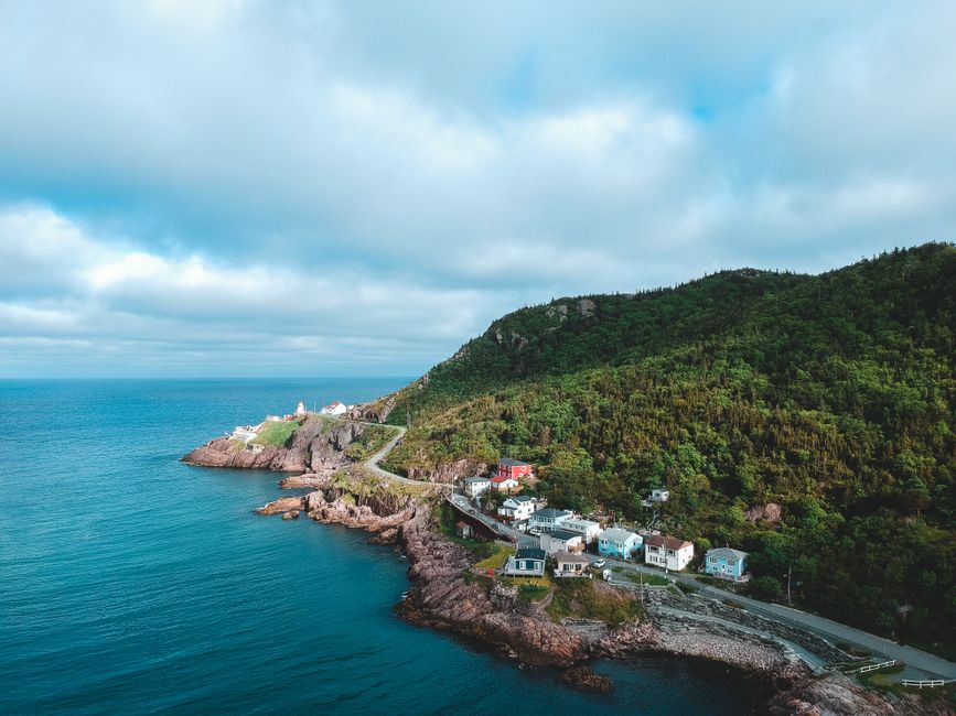 Aerial view of a coastal area showing a hillside covered in dense green foliage with residential houses positioned along a winding road close to the shoreline. The houses are small, with various colours including white, blue, and grey, and have pitched roofs. The rocky coastline features a narrow promenade or access path running alongside the water, with the sea appearing calm and blue-green. In the background, the sky is partly cloudy with patches of sunlight illuminating the landscape. The scene captures a serene seaside environment typical of a quiet coastal community, with no visible signs of rubbish or waste, emphasizing natural beauty and residential living near the water.