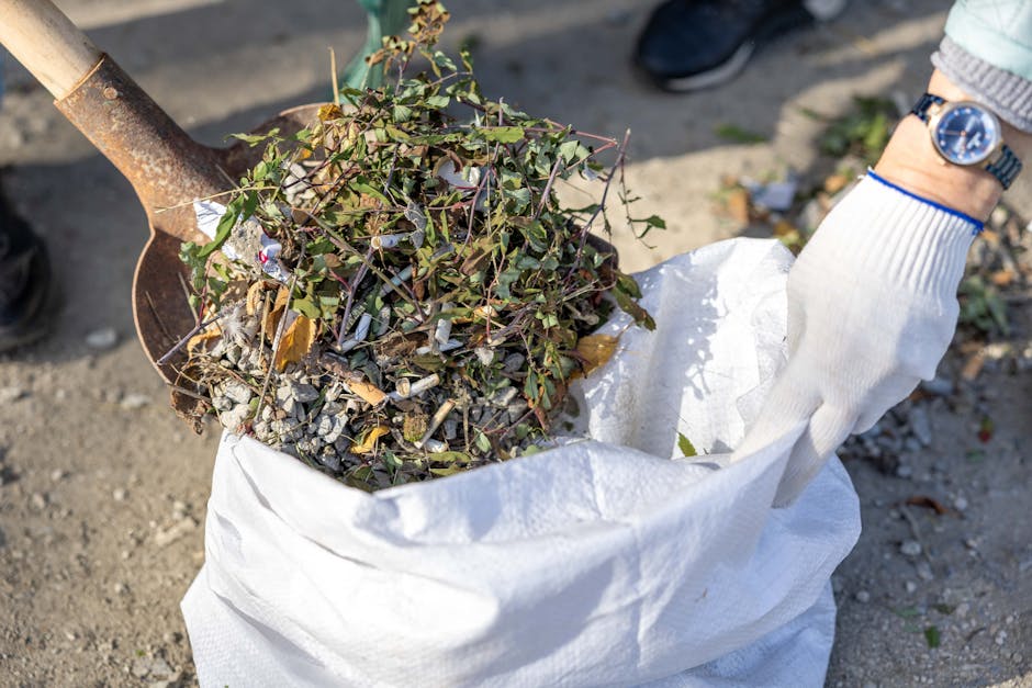 A person wearing a white glove and a wristwatch is holding a large white sack filled with garden debris, including intertwined green ivy, small branches, and scattered leaves. The sack, made of heavy-duty woven material, is placed on a concrete or asphalt surface, with loose gravel and small stones visible beneath. The scene appears outdoors, with natural lighting casting soft shadows on the ground and the discard material. A rusted shovel with a wooden handle is partially visible, leaning into the sack, indicating active debris collection or clearing. The background shows a portion of someone's dark footwear, suggesting a practical setting for garden waste removal. This image reflects an aspect of private or independent waste clearance, typical of rubbish removal services offered by companies such as wasteclearancestjohnswood.co.uk, emphasizing the gathering and disposal of garden rubbish as part of on-site clearance or alternative waste handling tasks.